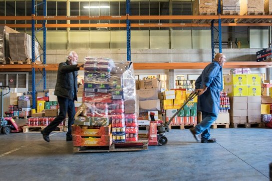 Twee oudere vrijwilligers van Fluvius – Goede doelen vervoeren voedselpakketten op een trekkar tussen de rekken in een groot magazijn van De Voedselbanken