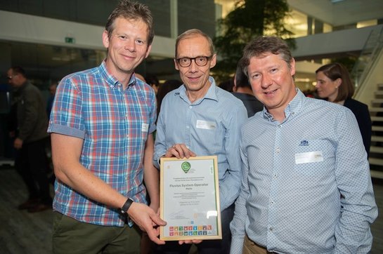 Uitreiking Voka Charter Duurzaam Ondernemen Drie mannen houden samen een houten kader met daarin het charter duurzaam ondernemen vast. Ze kijken fier en glimlachend in de camera.